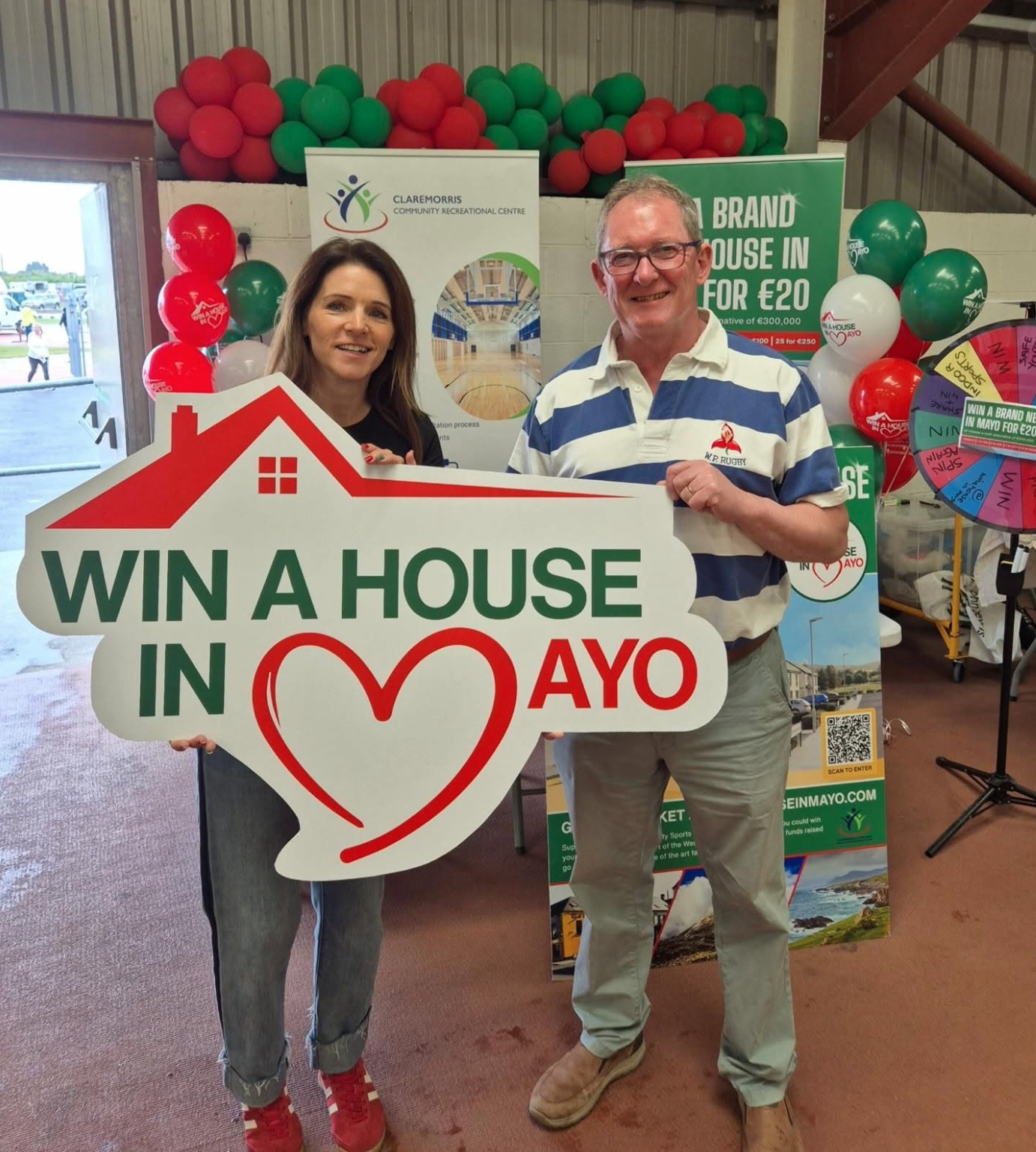 Two people smile holding a large sign reading “WIN A HOUSE IN MAYO” at an event.